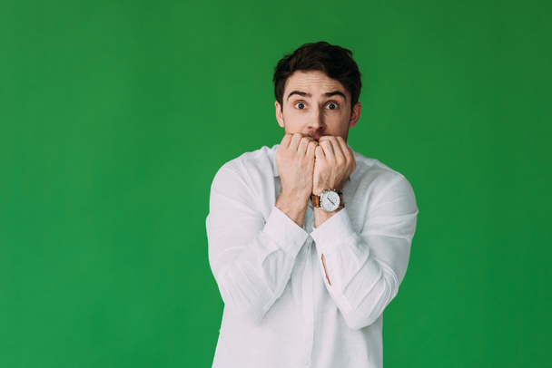 front view of scared young man in white shirt covering mouth with hands isolated on green - Фото, изображение