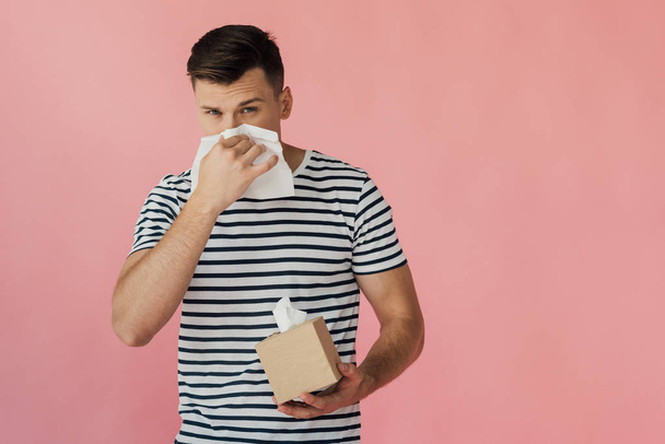 young man in striped t-shirt wiping nose with napkin isolated on pink - Фото, изображение