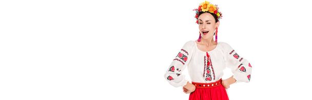 brunette jonge vrouw in nationaal Oekraïens kostuum met handen op heupen winking geïsoleerd op wit, panoramische schot - Foto, afbeelding
