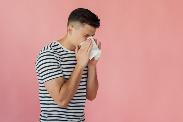 young man in striped t-shirt blowing nose and using napkin isolated on pink - Foto, imagen