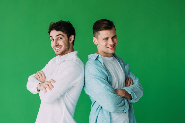 dwóch uśmiechniętych mężczyzn stojących z skrzyżowanymi ramionami i patrząc na aparat izolowany na zielono - Zdjęcie, obraz