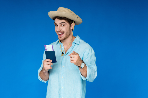 turista sonriente en sombrero de safari con pasaporte y billete de avión y apuntando con el dedo a la cámara aislado en azul
 - Foto, imagen
