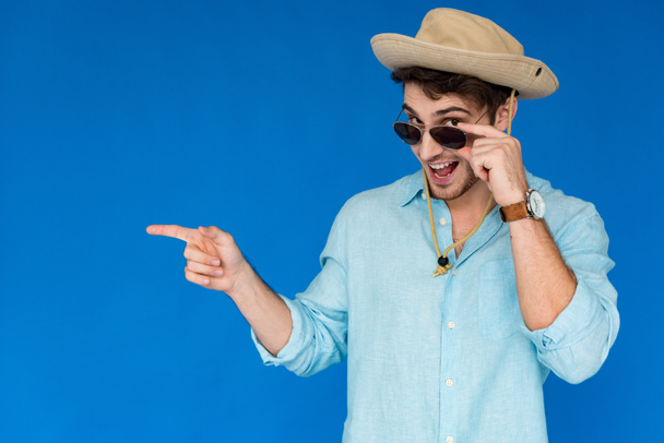 excited tourist in safari hat and sunglasses laughing and pointing with finger isolated on blue - Foto, immagini