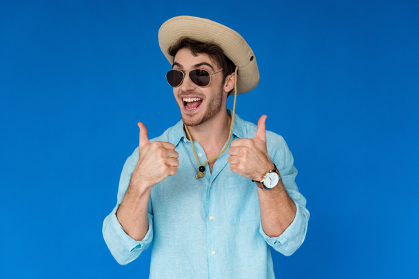 возбужденный путешественник в сафари шляпу и солнцезащитные очки, показывающие большие пальцы вверх изолированы на синий
 - Фото, изображение