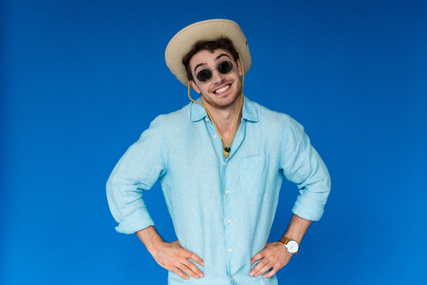 front view of smiling traveler in safari hat and sunglasses standing with hands on hips isolated on blue - Φωτογραφία, εικόνα