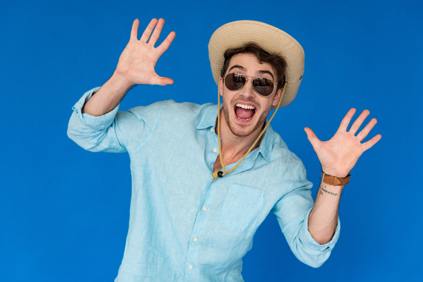 front view of excited traveler in safari hat and sunglasses standing with hands up isolated on blue - Φωτογραφία, εικόνα
