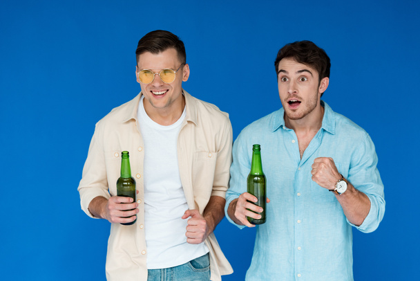 δύο φίλοι που κρατούν μπουκάλια μπύρας και κοιτάζουν μακριά απομονωμένοι στο μπλε - Φωτογραφία, εικόνα