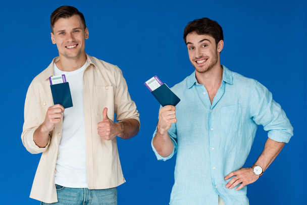 two smiling tourists holding passports with air tickets and showing thumb up isolated on blue - Photo, Image