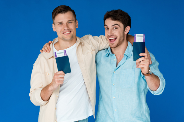 two smiling tourists holding passports with air tickets and embracing isolated on blue - Photo, Image