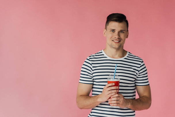 front view of smiling young man in striped t-shirt holding beverage isolated on pink - Photo, Image