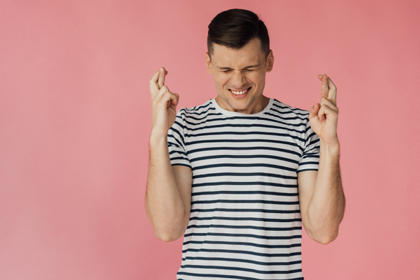 front view of worried young man in striped t-shirt with crossed fingers isolated on pink - Foto, Imagem