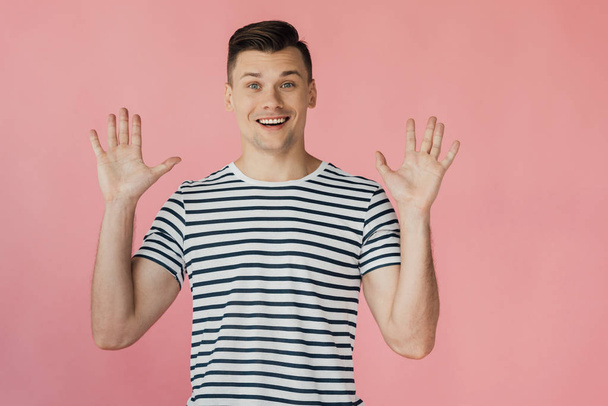 front view of excited smiling young man in striped t-shirt standing with hands up isolated on pink - Photo, image