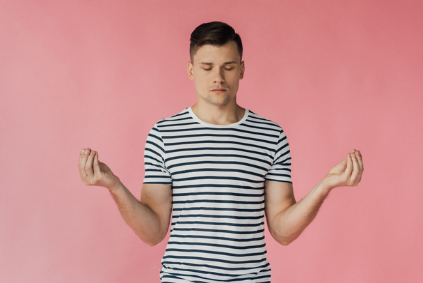 front view of relaxed young man in striped t-shirt meditating with closed eyes isolated on pink - Foto, imagen