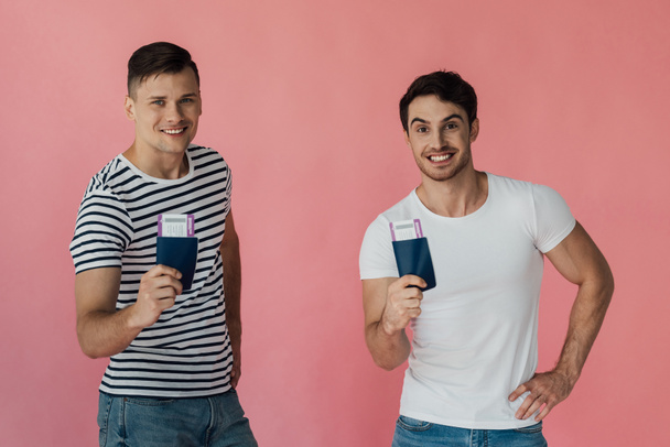 vista frontal de dos turistas emocionados sonriendo y sosteniendo pasaportes con billetes de avión aislados en rosa
 - Foto, imagen