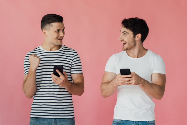 dos hombres sonrientes usando teléfonos inteligentes y mirándose aislados en rosa
 - Foto, imagen