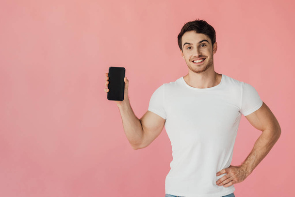 front view of laughing muscular man in white t-shirt standing with hand on hip and holding smartphone with blank screen isolated on pink - Fotó, kép
