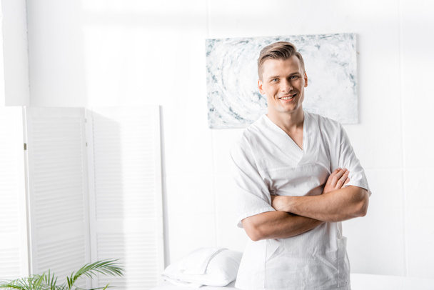 smiling masseur standing with crossed arms and looking at camera in clinic - Fotó, kép