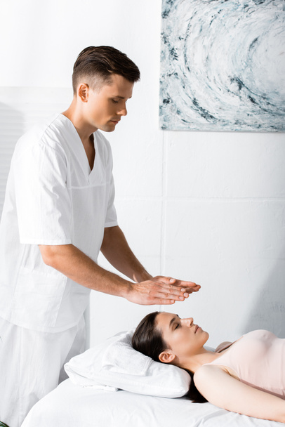 focado curandeiro de pé perto da mulher na mesa de massagem e de mãos dadas acima de sua cabeça
 - Foto, Imagem