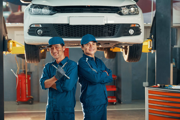 Портрет двох щасливих автомеханіків в уніформі, що працюють в автосервісі з автомобілем на задньому плані
  - Фото, зображення