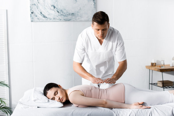 focused healer standing near woman lying on massage table with closed eyes and holding hands above her body - Photo, Image