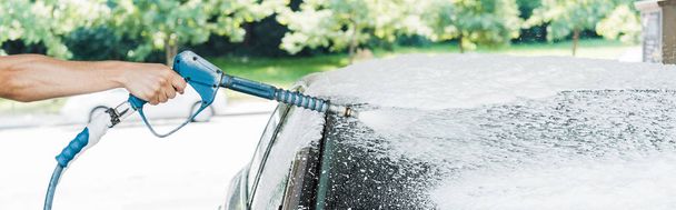 panoramic shot of man holding pressure washer with foam near car  - Foto, imagen