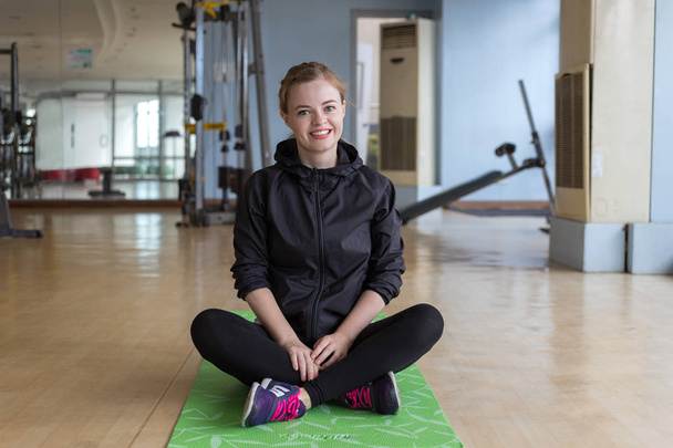 Joven mujer caucásica sentada en una alfombra en el gimnasio sonriendo
 - Foto, Imagen