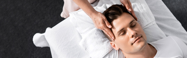 tiro panorâmico de massagista em pé perto do homem com os olhos fechados e tocando seu rosto
 - Foto, Imagem