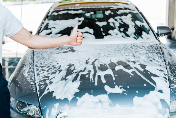 cropped view of car cleaner showing thumb up near car with foam  - Foto, afbeelding