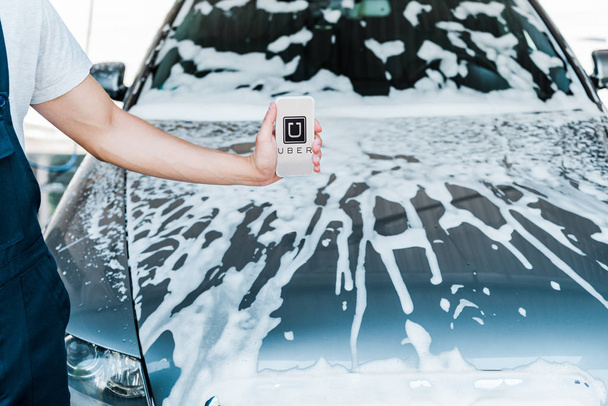 KYIV, UKRAINE - JUNE 20, 2019: cropped view of man holding smartphone with uber app on screen near car with foam  - 写真・画像
