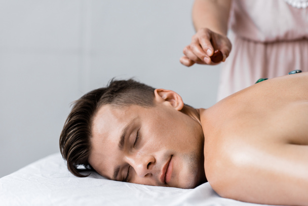 bijgesneden beeld van masseur en shirtless man met stenen op rug - Foto, afbeelding
