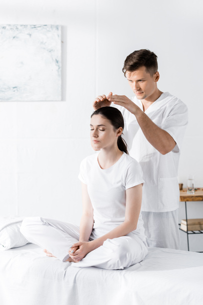 woman sitting on massage table with closed eyes while healer holding hands above her head - Foto, afbeelding