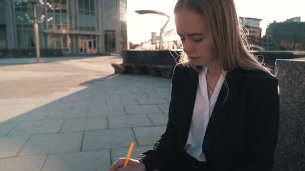 Caucasian young business woman in white shirt sits on a bench on the street and works. The girl writes in a notebook - Materiał filmowy, wideo