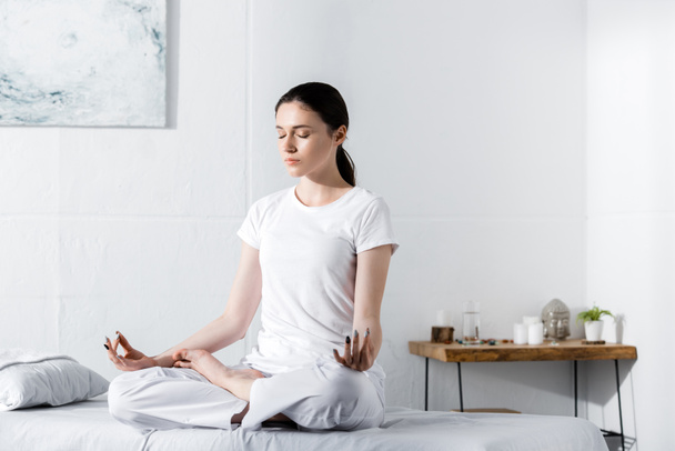 young woman sitting in lotus pose with closed eyes on massage table - Photo, Image