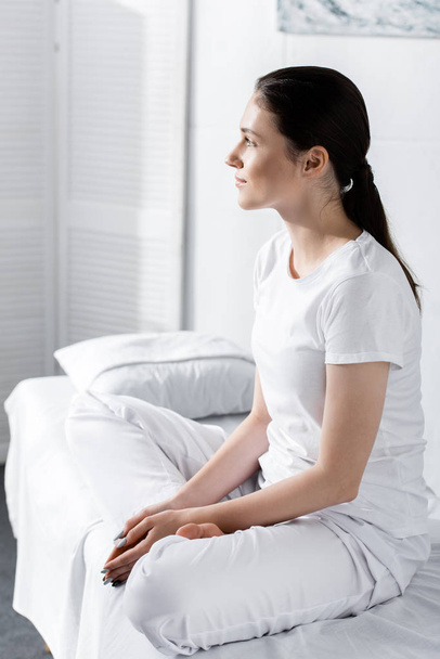attractive young woman with ponytail sitting on bed and looking away in clinic - Foto, Imagen
