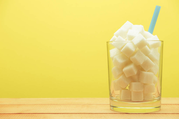 Glass full of cubes sugar with straw on a wooden table  beautiful yellow  background  - unhealthy diet concept. - Fotografie, Obrázek
