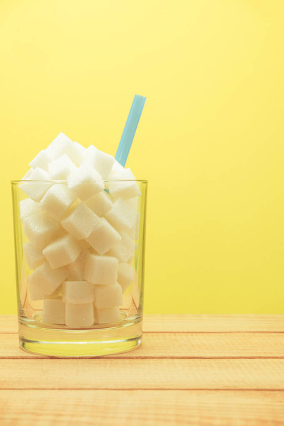 Glass full of cubes sugar with straw on a wooden table  beautiful yellow  background  - unhealthy diet concept. - Foto, Imagen