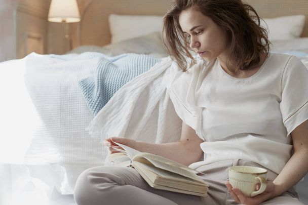 Молода жінка сидить біля ліжка і читає книгу
 - Фото, зображення