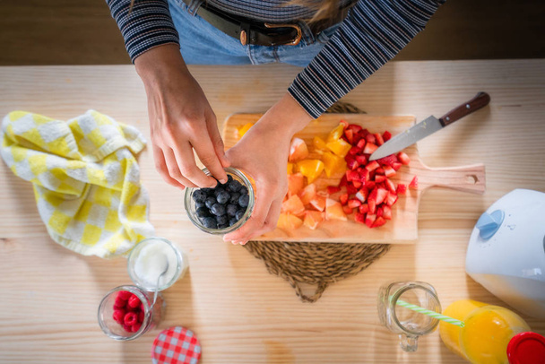 geschnittenes Bild einer Frau, die Blaubeeren aus dem Glas nimmt, während sie zu Hause gesunde Vitaminnahrung aus frischen Früchten kocht - Foto, Bild