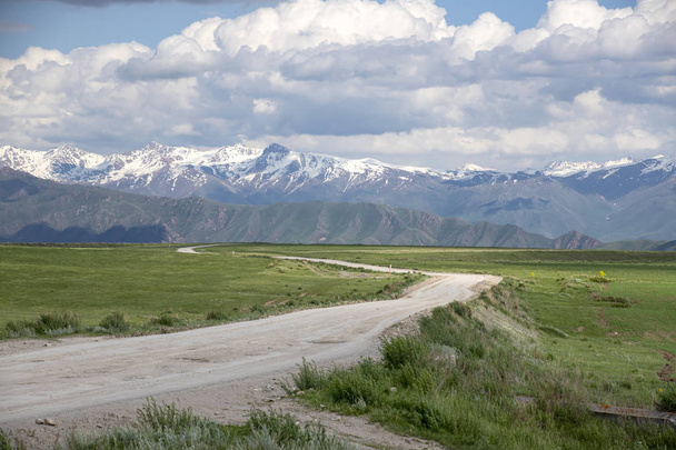 Country road among green fields and hills against the backdrop of mountains with snow-capped peaks - Photo, Image
