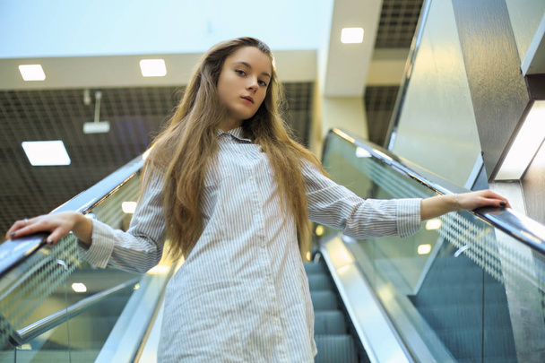 Portrait of a young pretty caucasian teenage girl with long hair in a shirt on the escalator in a mall. - Foto, imagen