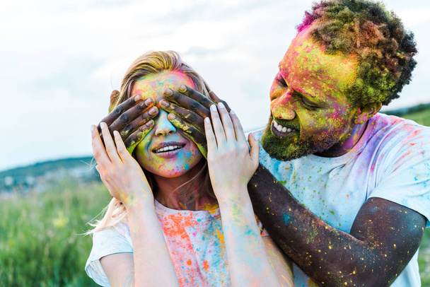happy african american man covering eyes of young woman with holi paints on face  - Φωτογραφία, εικόνα