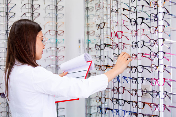 Приваблива жінка-офтальмолог пише дані в буфер обміну, працює в оптичному магазині. Концепція охорони здоров'я та медицини
 - Фото, зображення