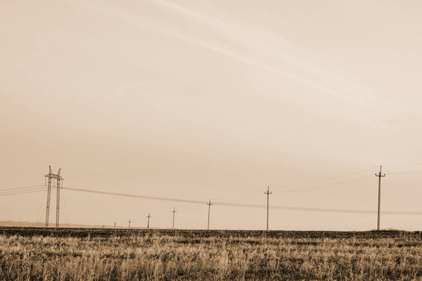 Atmospheric landscape with power lines in field under sky in sepia tones. Background image of electric pillars with copy space. Wires of high voltage above ground. Electricity industry in monochrome. - Фото, изображение