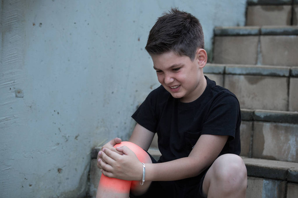 Junge mit Schmerzen, der auf sein verletztes Bein blickt, das auf der Treppe sitzt. Knieschmerzen - Foto, Bild