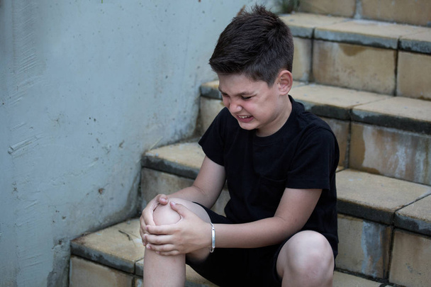 痛みを感じている少年は、負傷した脚を階段に座って見ている。膝の痛み - 写真・画像