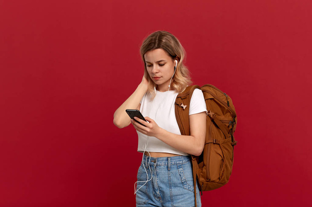 Portret dziewczyny z Kręcone blond włosy ubrane w białym t-shirt stojący na czerwonym tle. Szczęśliwy model z pomarańczowym plecakiem i białym zestawem słuchawkowym patrzy na ekran telefonu. Koncepcja podróżowania - Zdjęcie, obraz