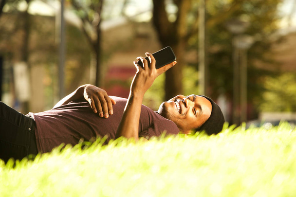 Portrét šťastného afroamerického muže relaxujícího v parku a pohledu na mobilní telefon - Fotografie, Obrázek