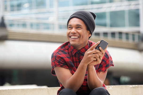 Portret van gelukkige jonge zwarte man met muts zittend buiten in de stad met mobiele telefoon en op zoek weg - Foto, afbeelding