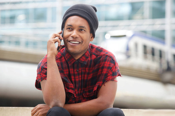 Portrait of cool young black guy sitting outside in city and talking with cellphone - Фото, изображение