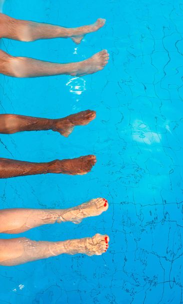 ноги трех друзей (простых людей) разных рас, сидящих у бассейна в солнечный день. Вечеринка Саммер. Отдых, международная дружба и спортивная концепция
. - Фото, изображение